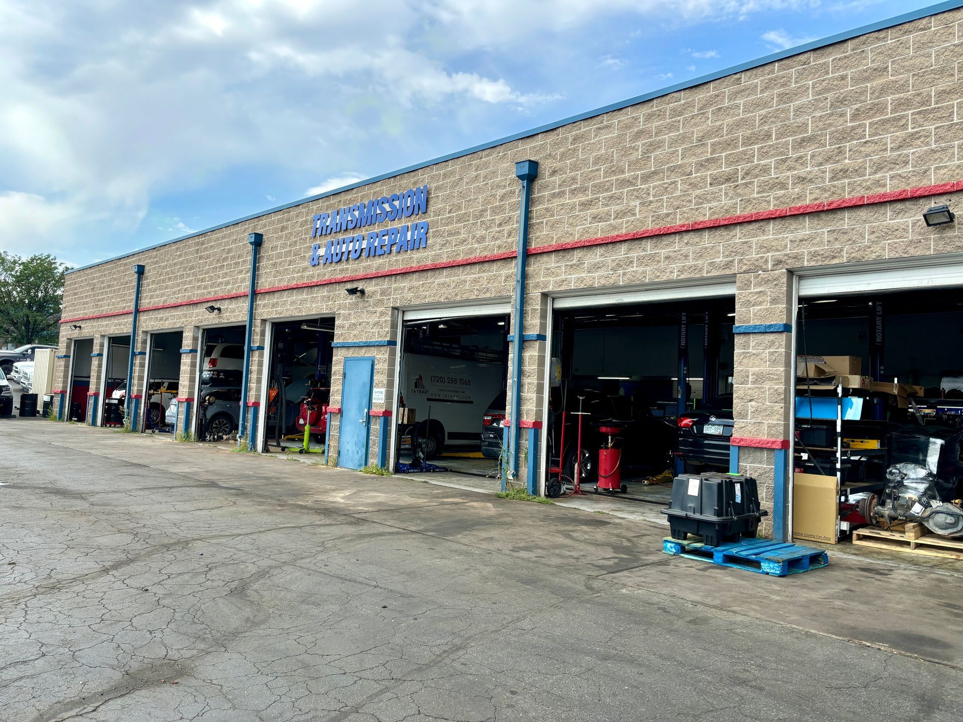 Exterior of a brick auto repair shop with several open garage bays, parked cars, and equipment visible.
