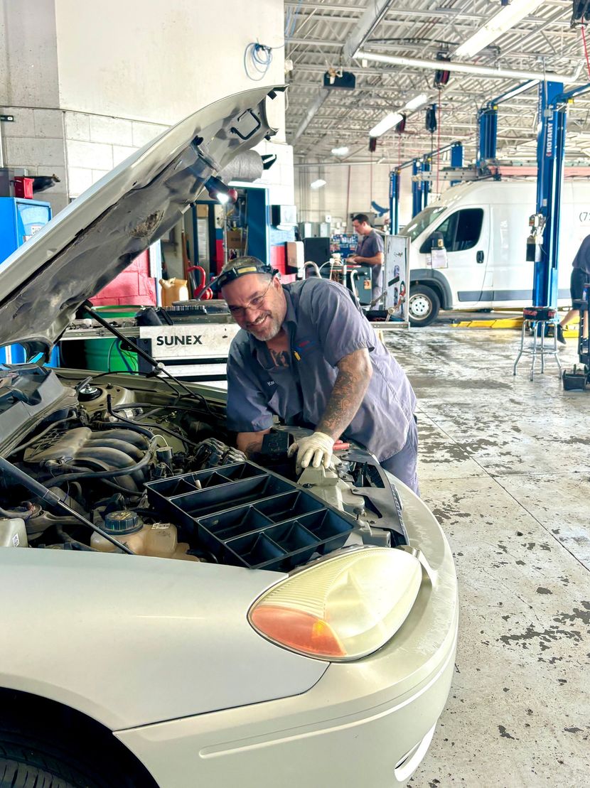 Mechanic working on a silver car with open hood in a garage. He smiles. Blue lift posts in background.