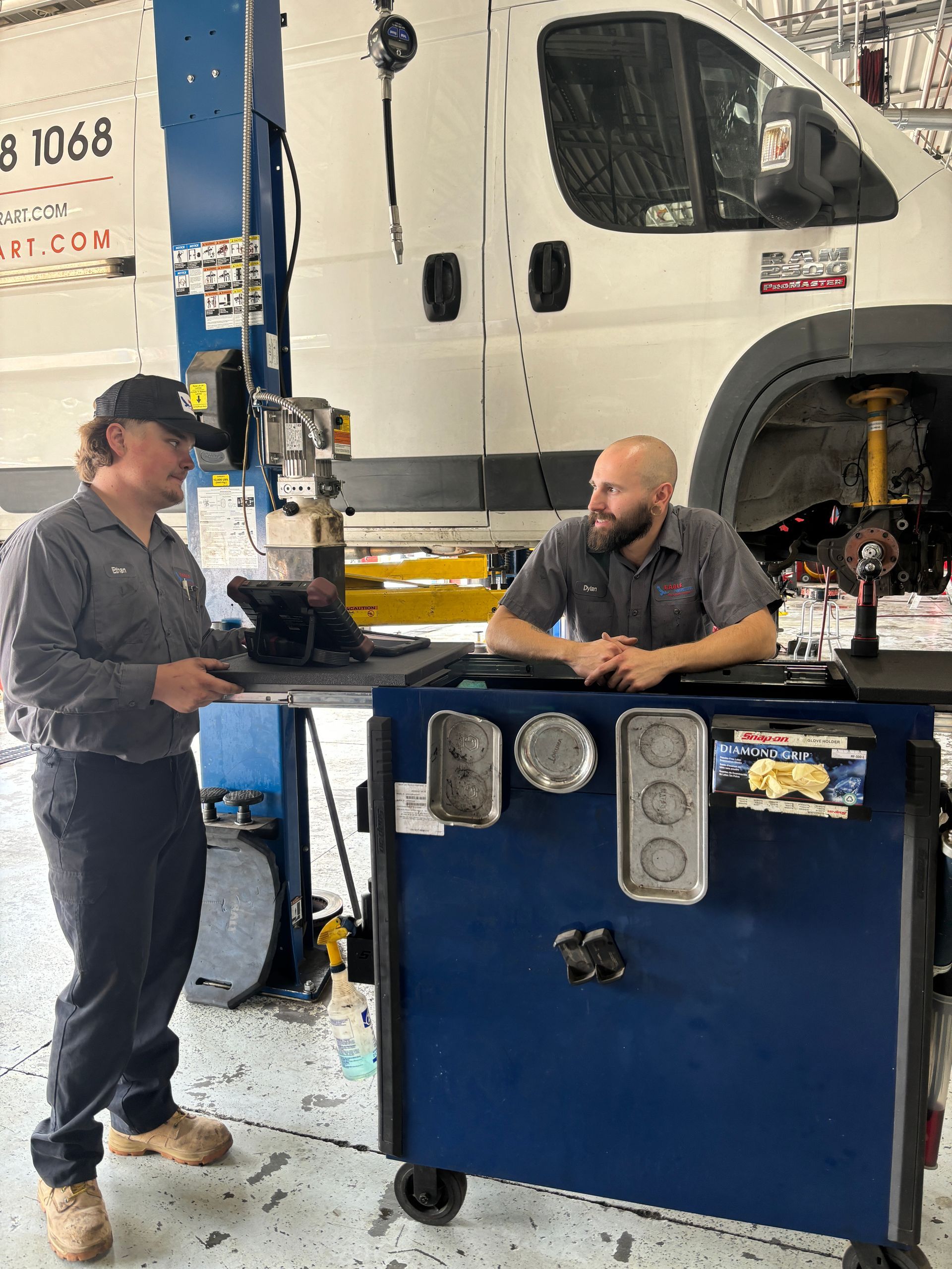 Two mechanics conversing in a garage. One holds a part, while the other leans on a toolbox near a raised van.