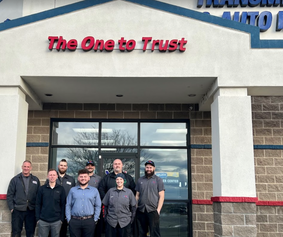 Group of auto repair shop employees smiling in front of the business entrance. Sign reads 