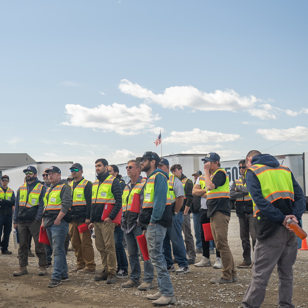 A group of workers in safety vests stand outdoors under a cloudy sky.