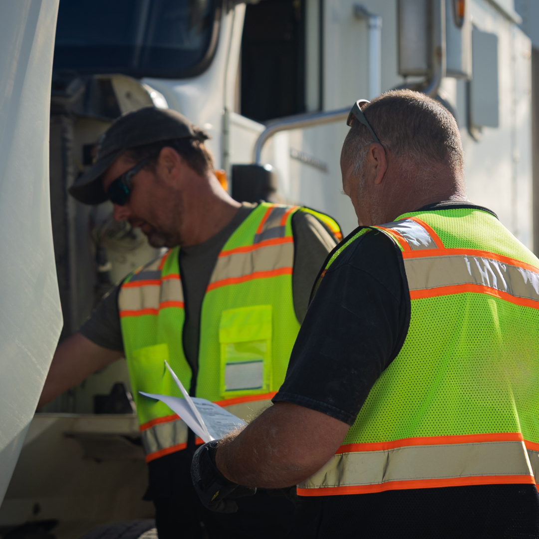 Two people in safety vests reviewing paperwork near a truck.