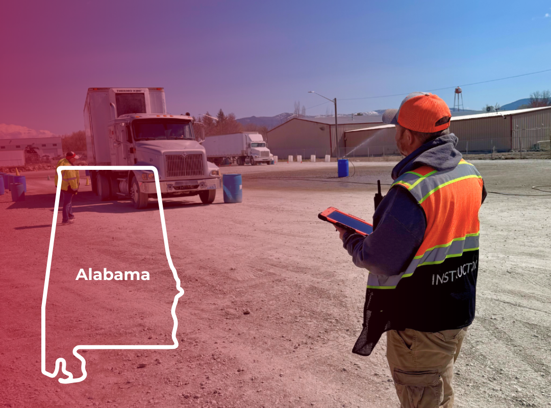 Inspector in orange vest and hard hat observes a truck. Another worker stands by the truck. Outdoor setting.