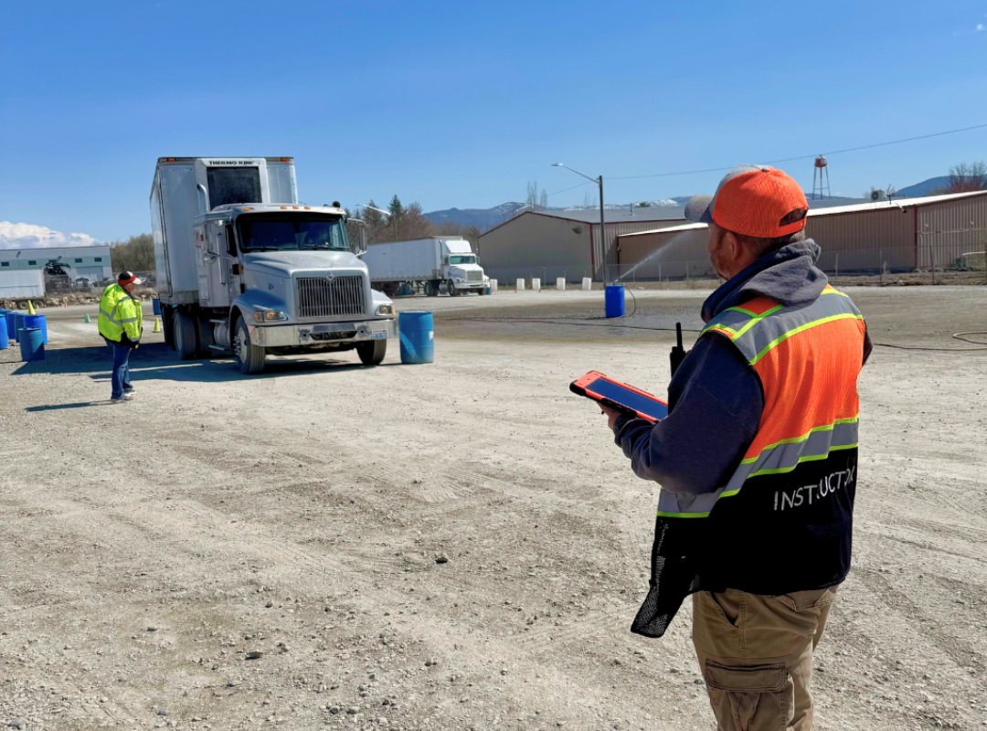 Inspector in orange vest and hard hat observes a truck. Another worker stands by the truck. Outdoor setting.