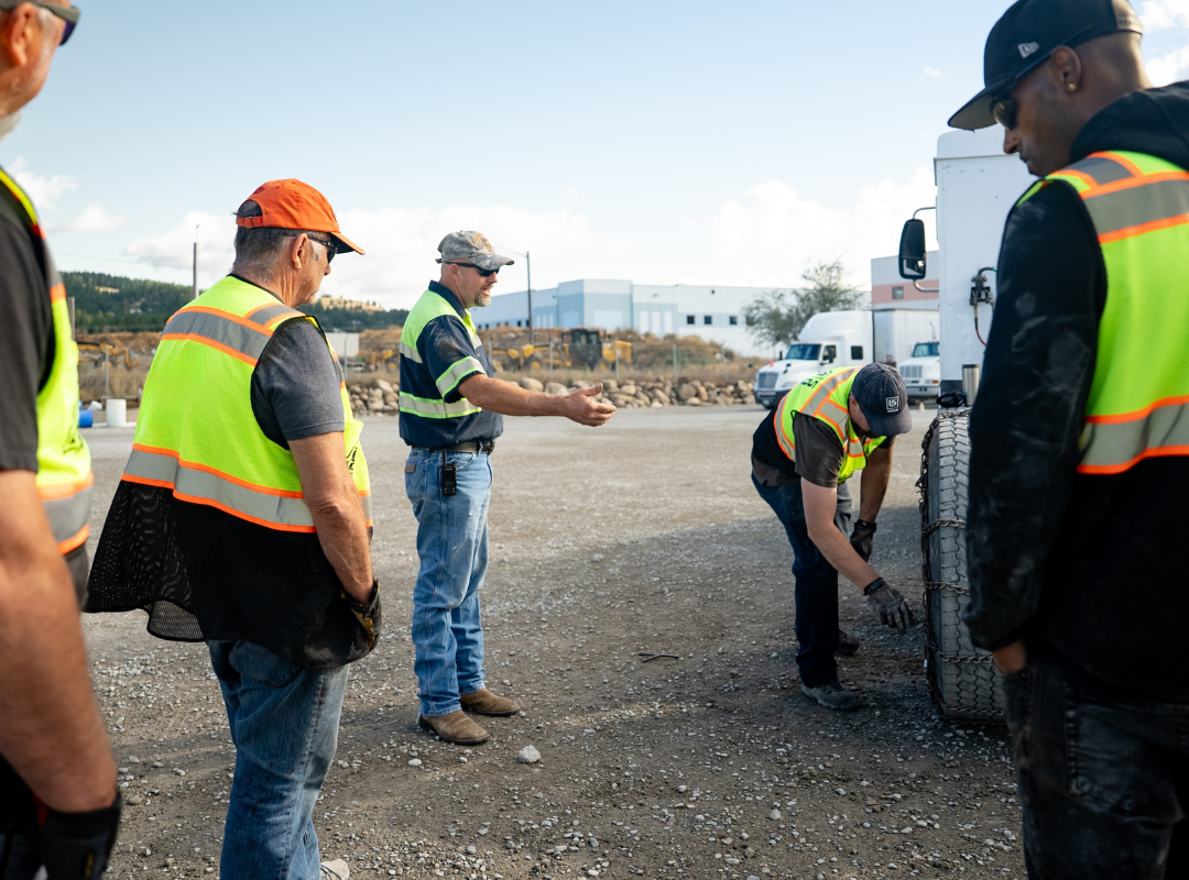 Men in reflective vests inspecting a truck tire in a gravel lot.