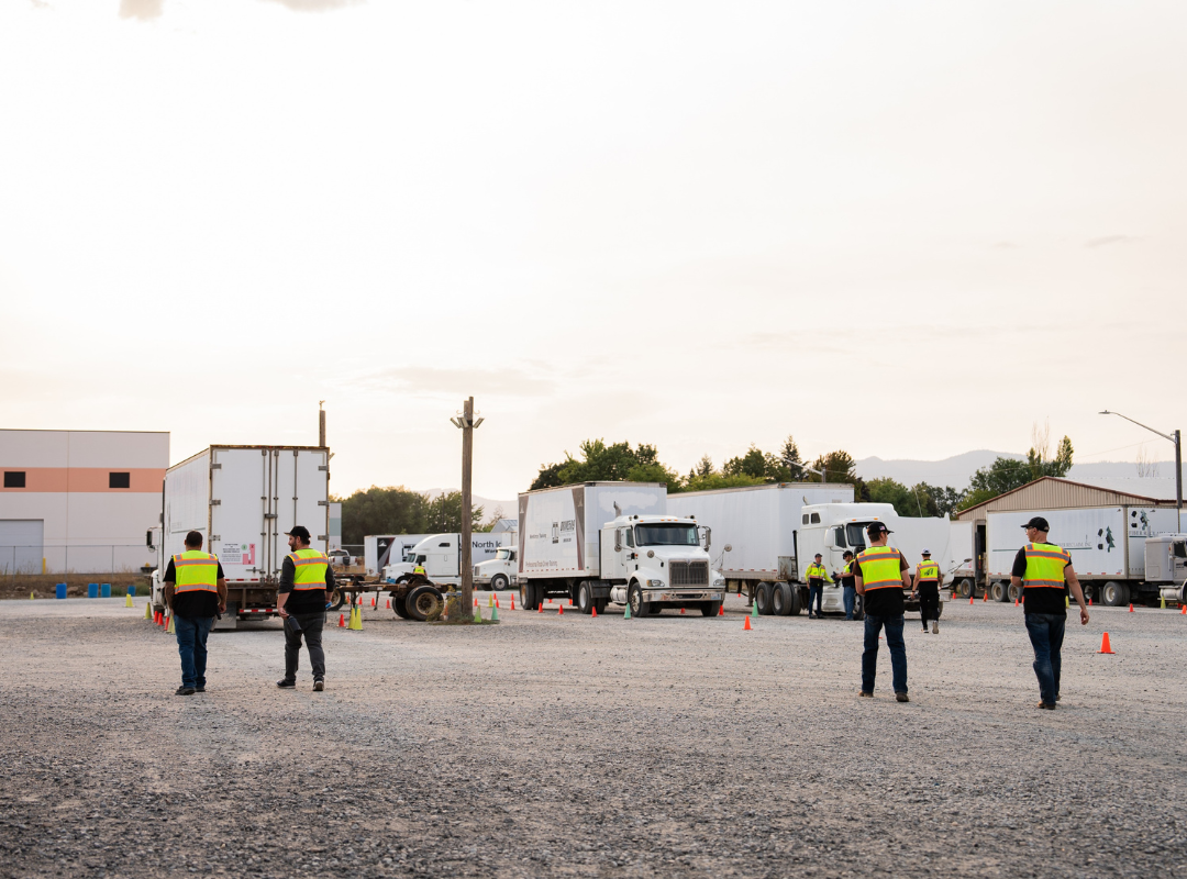 Two cdl students in high-visibility vests work on the landing gear of a semi-truck trailer outdoors.