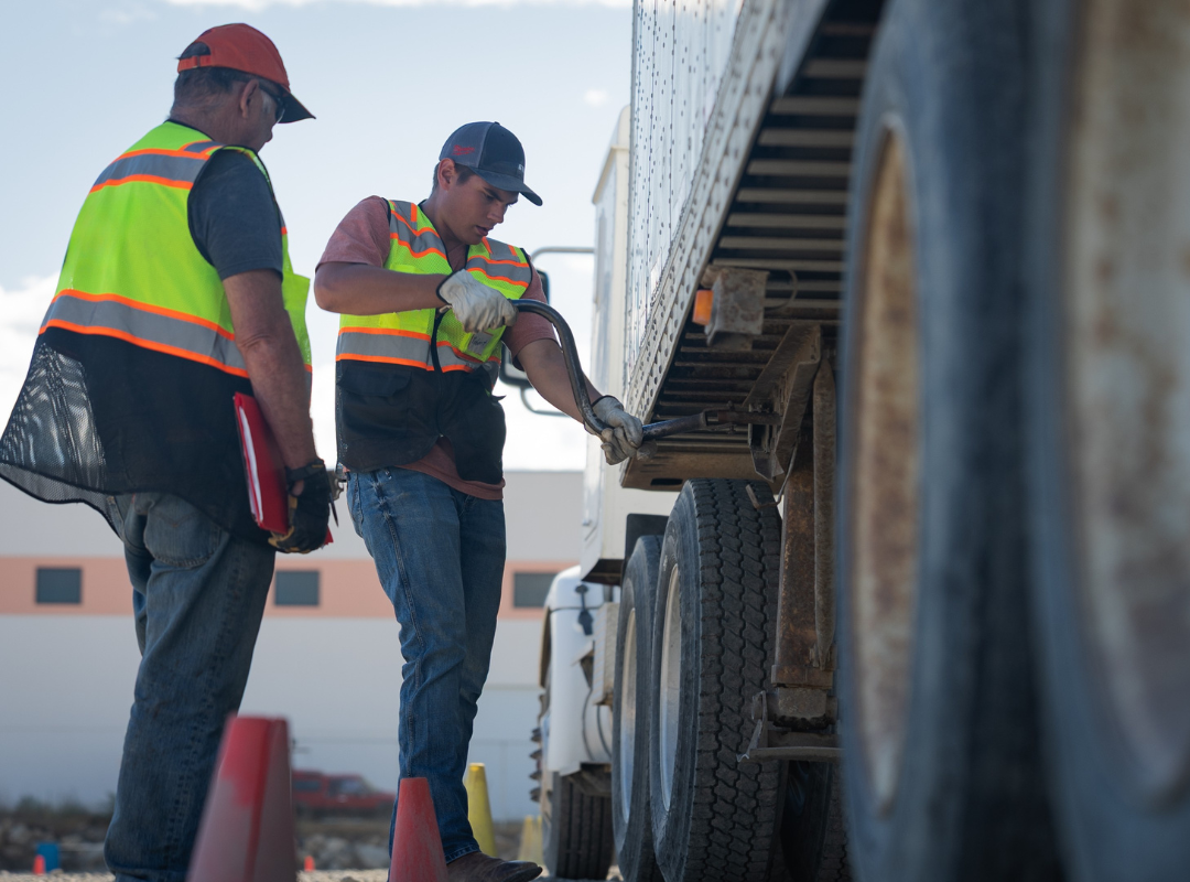 Two people in safety vests inspect a truck's tires outdoors on a sunny day.