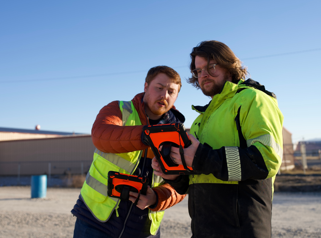 Two cdl instructors outdoors examining tablets, wearing safety vests. One points, discussing cdl training compliance.