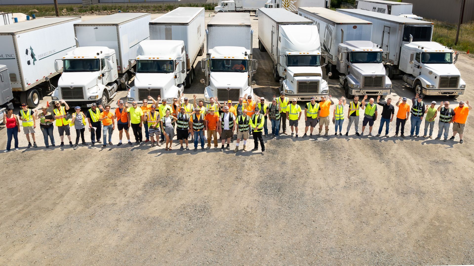 A large group of people in high-visibility vests stands in a line in front of several semi-trucks in a gravel lot.