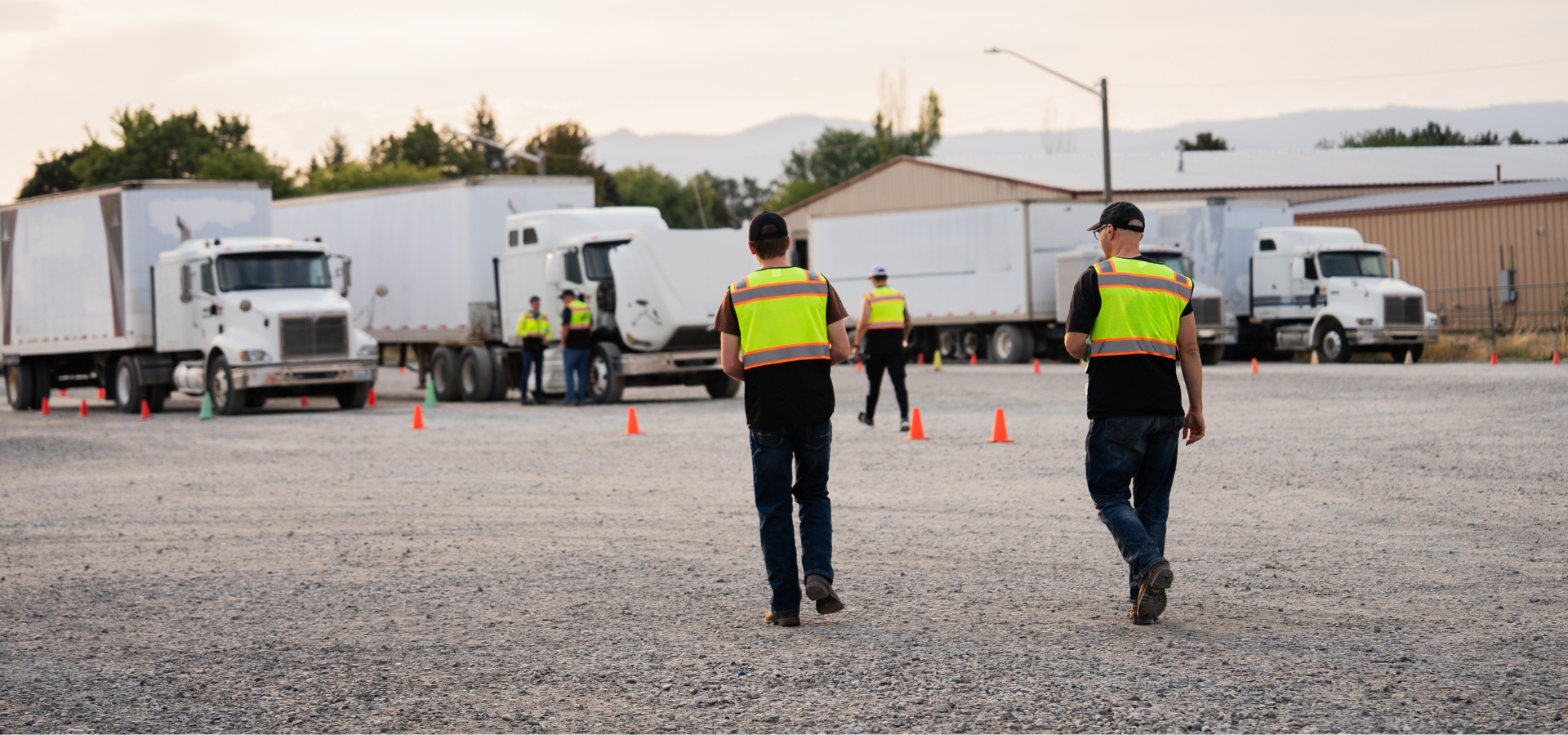 Truck yard with workers in yellow vests, trailers, and a warehouse at sunset