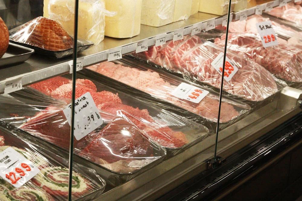 A refrigerated display case in a grocery store featuring various cuts of raw meat and blocks of cheese.