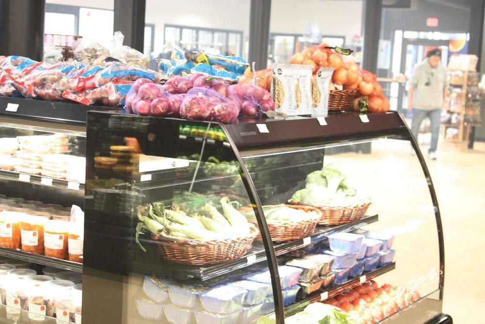A refrigerated grocery display case stocked with fresh produce like corn, onions, and oranges, with a person walking by.