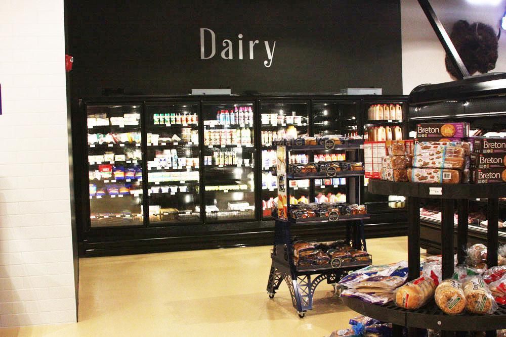 A grocery store dairy section with black refrigerated cases and a display rack of packaged bread goods in the foreground.