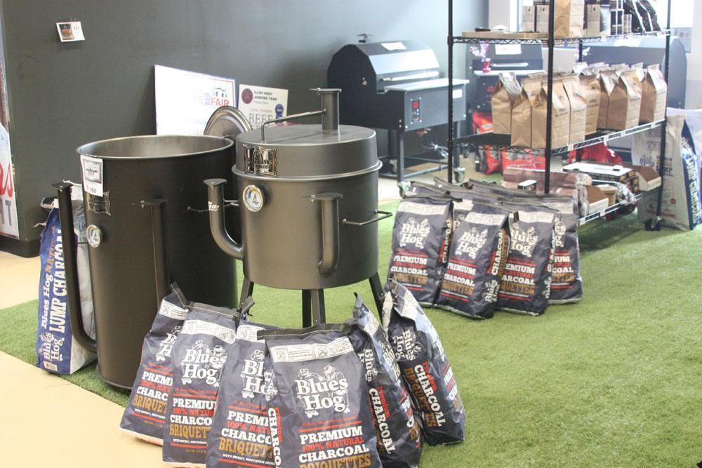 Black drum smokers and bags of Blues Hog charcoal on a green turf floor inside a retail store.