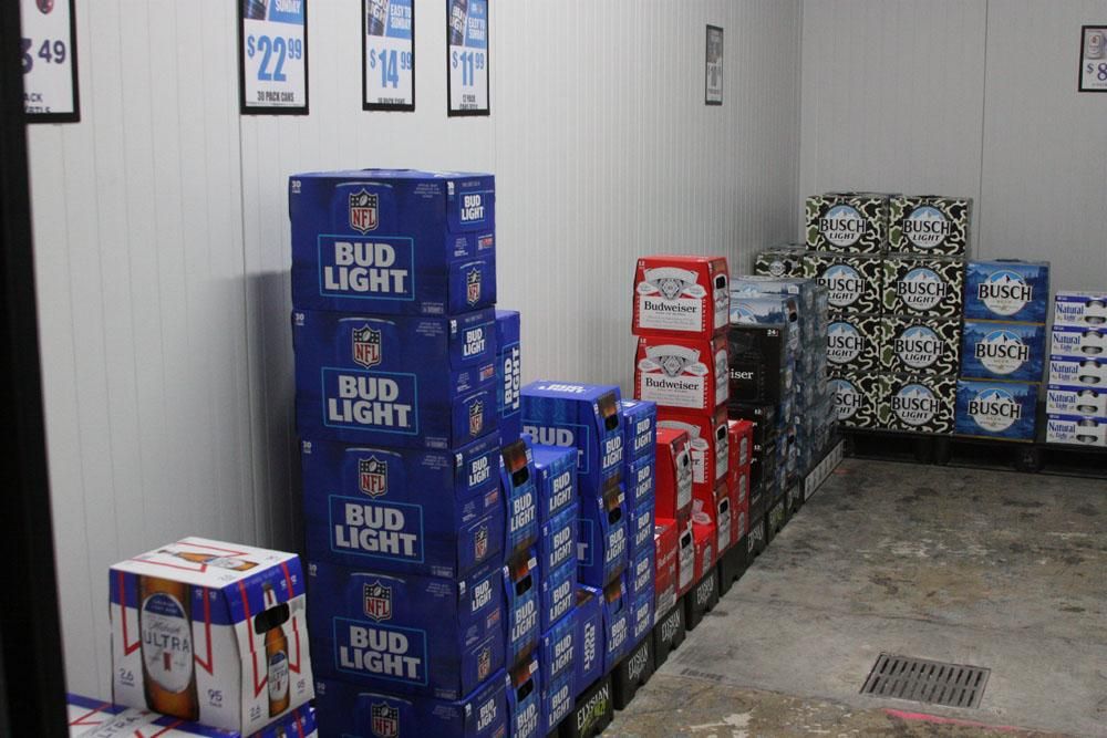 Stacked cases of Bud Light, Budweiser, and other beer brands stored in a refrigerated walk-in retail cooler.