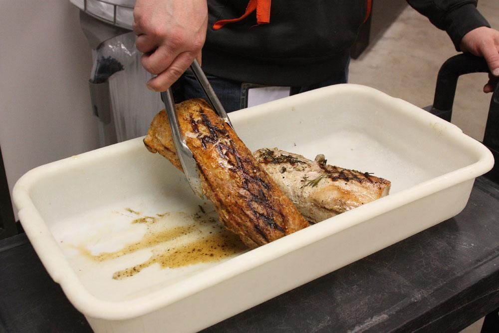 A pair of tongs holds a seared, grilled meat loin over a white plastic food service container next to a second loin.