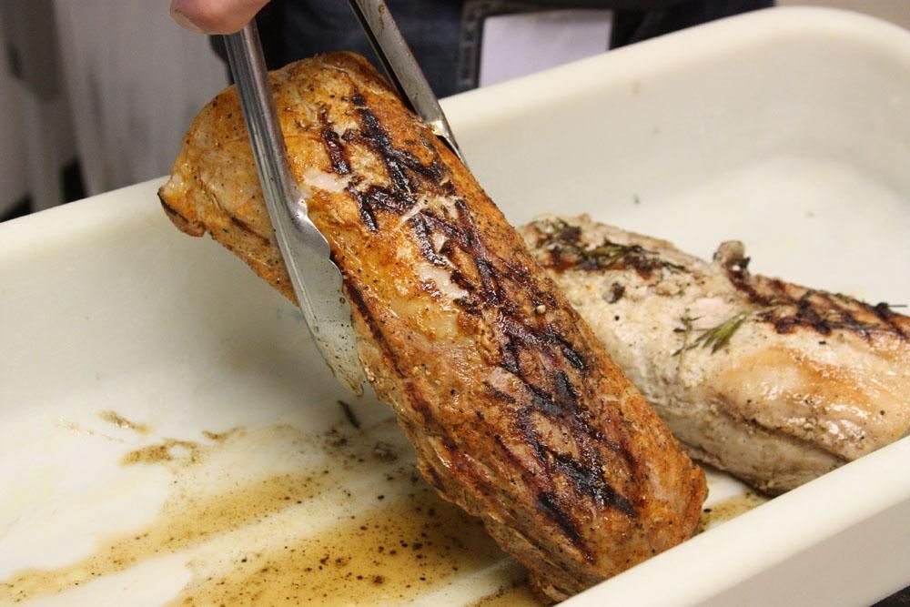 A person uses metal tongs to lift a piece of grilled, seasoned pork tenderloin from a white rectangular tray.