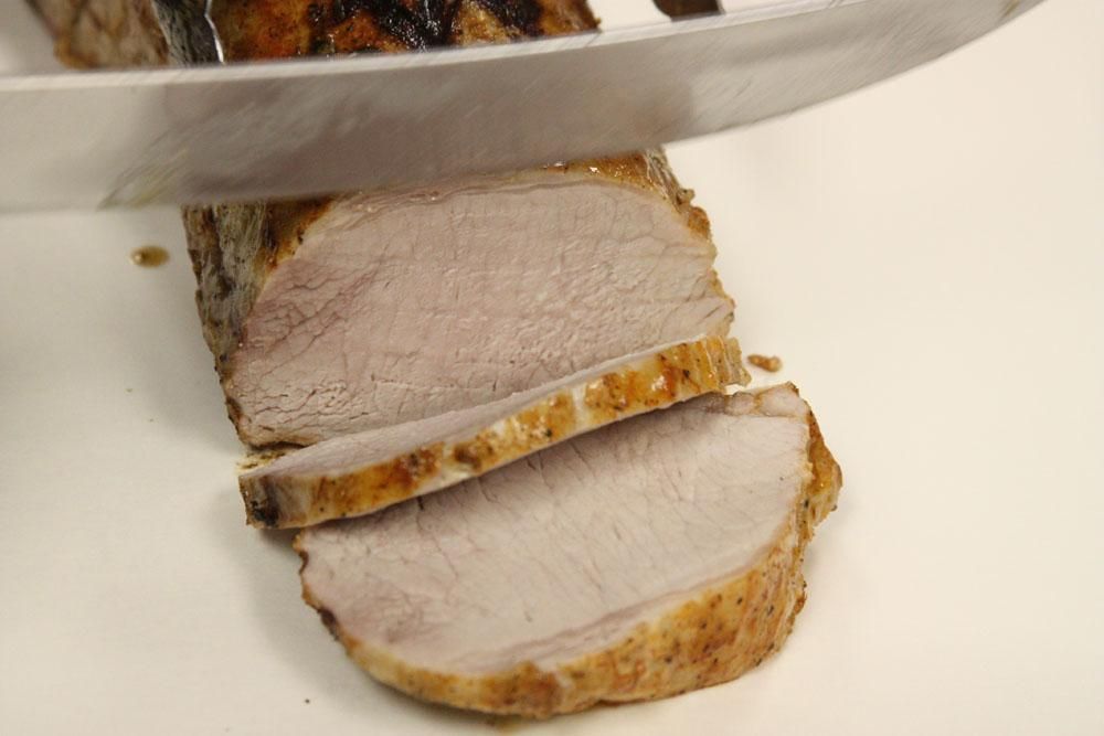 A knife cuts into a cooked pork tenderloin, showing sliced, light brown meat on a white surface.