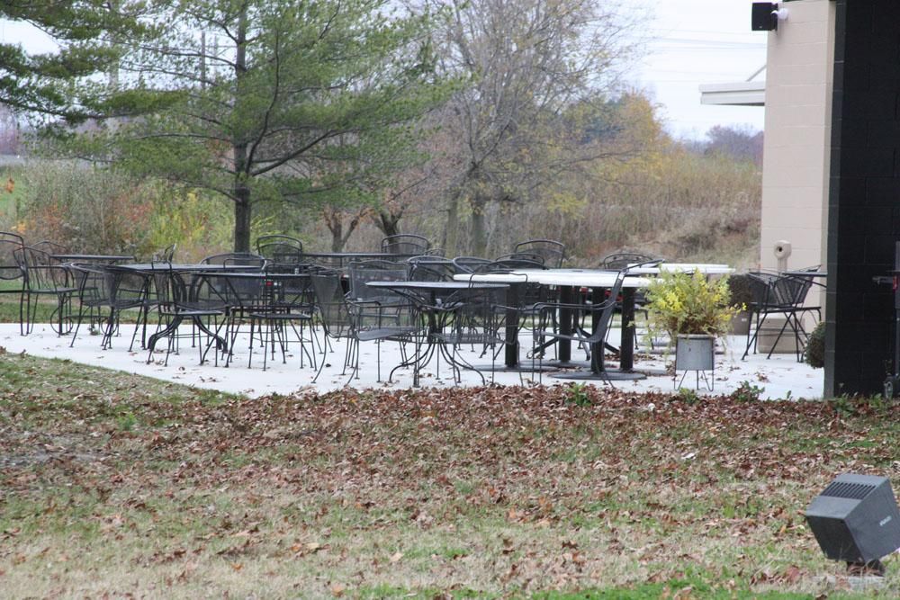 An outdoor patio area with multiple tables and chairs on a paved surface, surrounded by trees and dry grass.