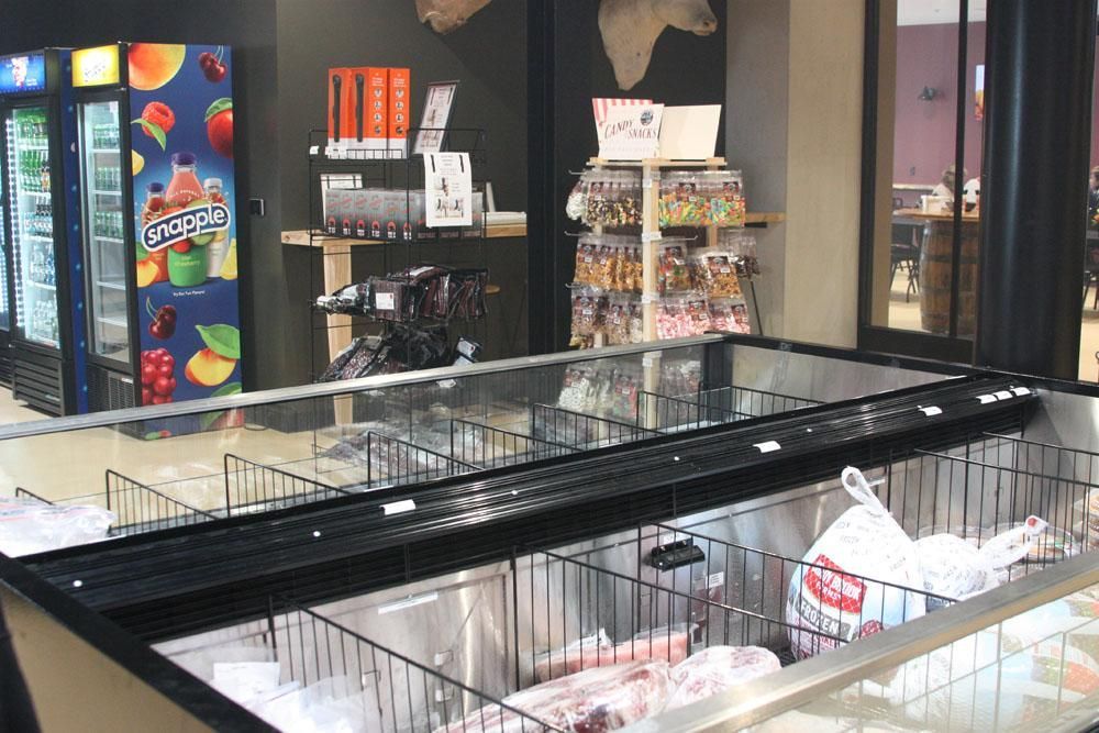 A grocery store display freezer containing frozen goods, with a refrigerated drink cooler and a snack stand in the back.