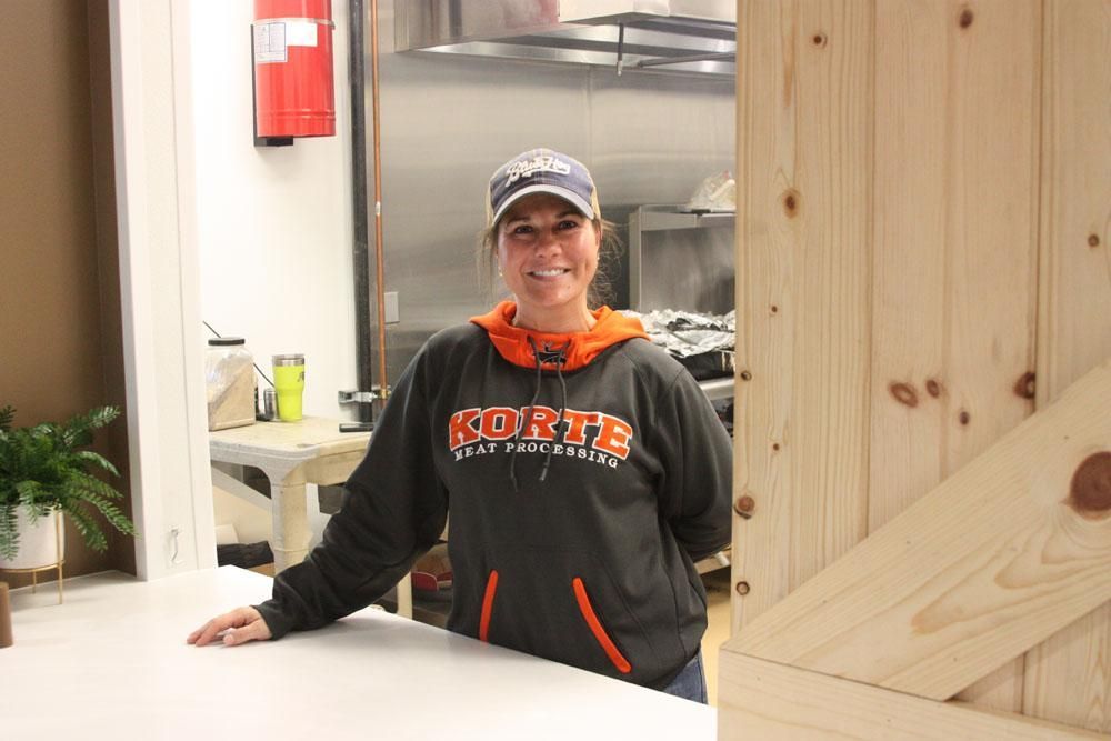 A person in a gray and orange Korte hoodie and a baseball cap smiling while standing behind a counter in a kitchen.