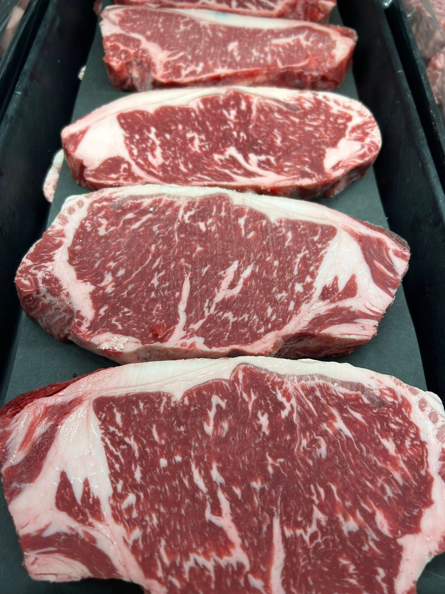 Five raw, marbled beef strip steaks arranged in a vertical row on a black surface.