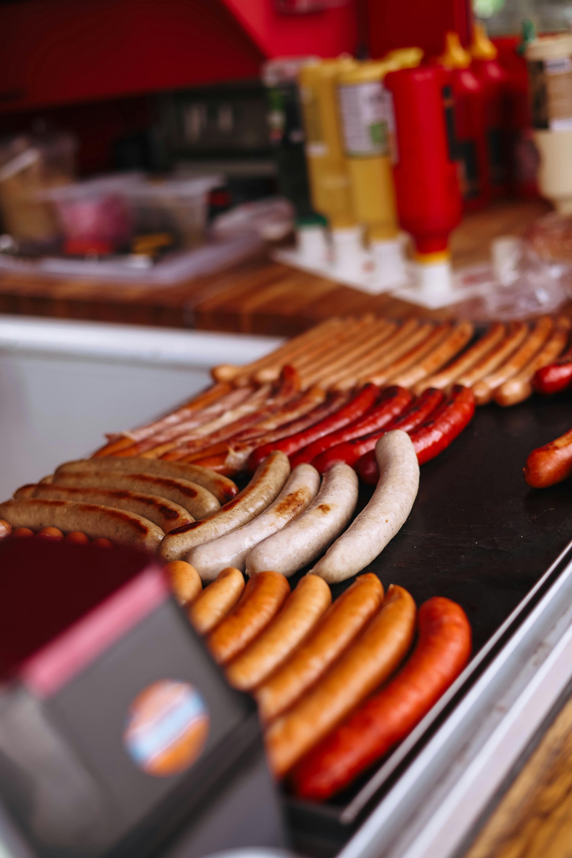 A close-up view of various types of sausages sizzling on a grill, with condiment bottles in the blurred background.