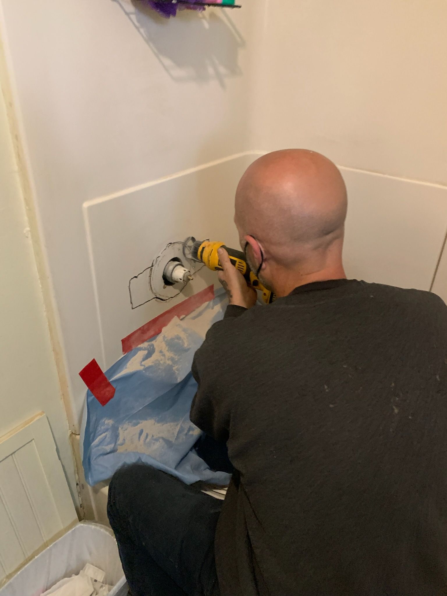 Person kneeling and drilling a wall fixture in a bathroom, with plastic sheeting covering the floor.