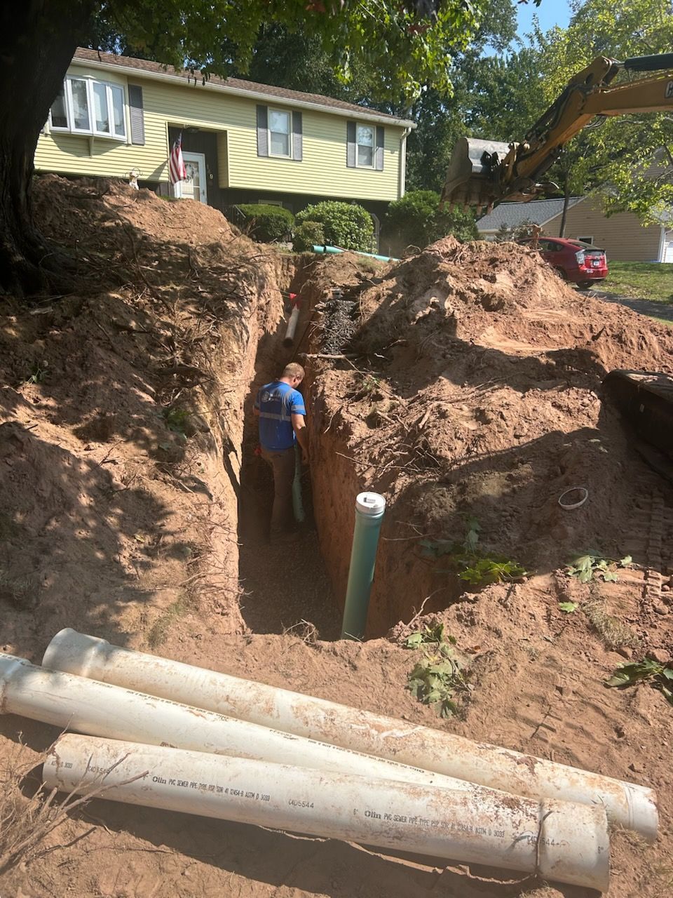 Worker standing in a deep excavation beside exposed pipes below a house hillside yard