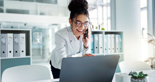 Woman in white blazer smiling on a phone call beside a laptop in a bright office Woman in white blazer smiling on a phone call beside a laptop in a bright office