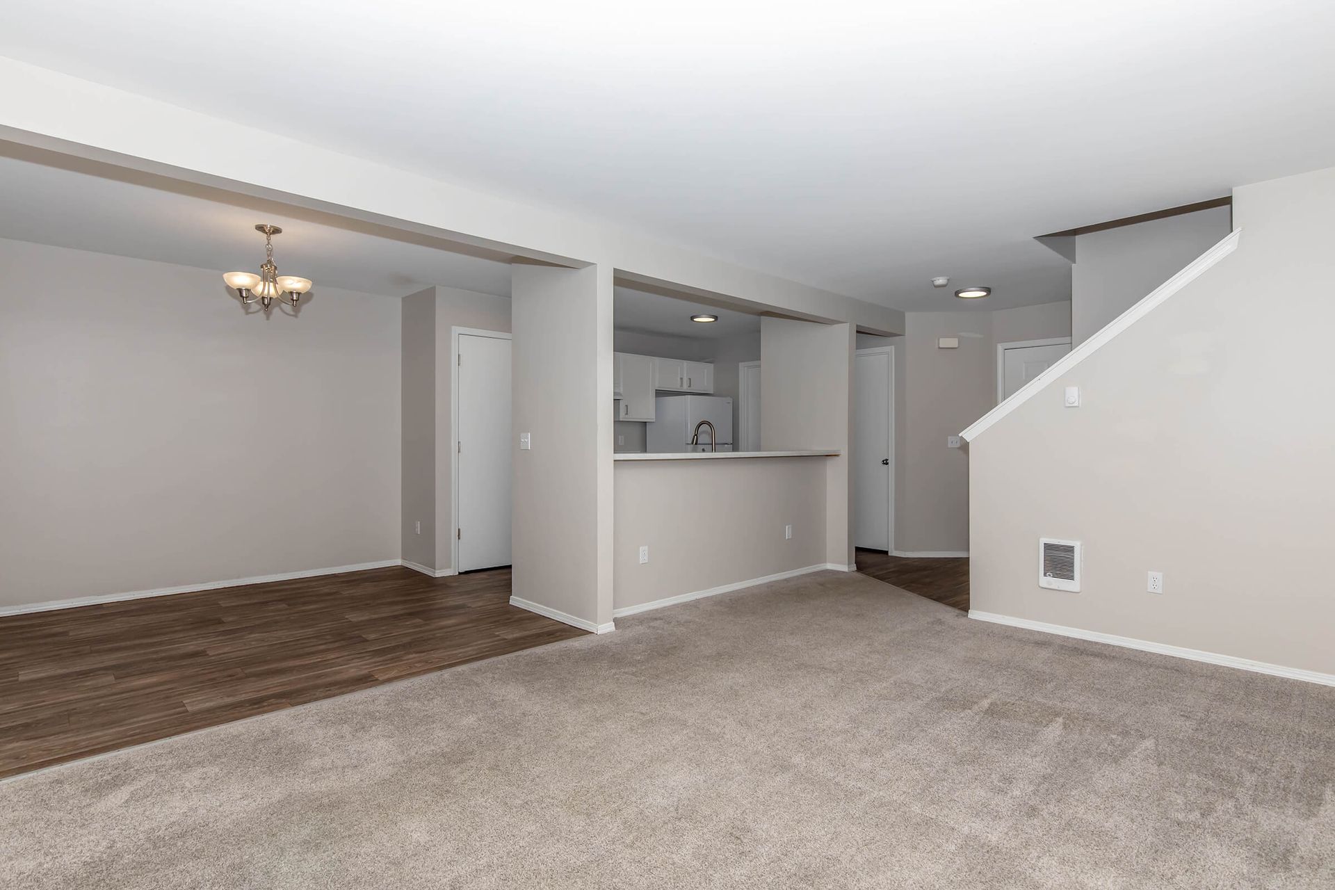 An empty living room with a carpeted floor and a kitchen in the background.