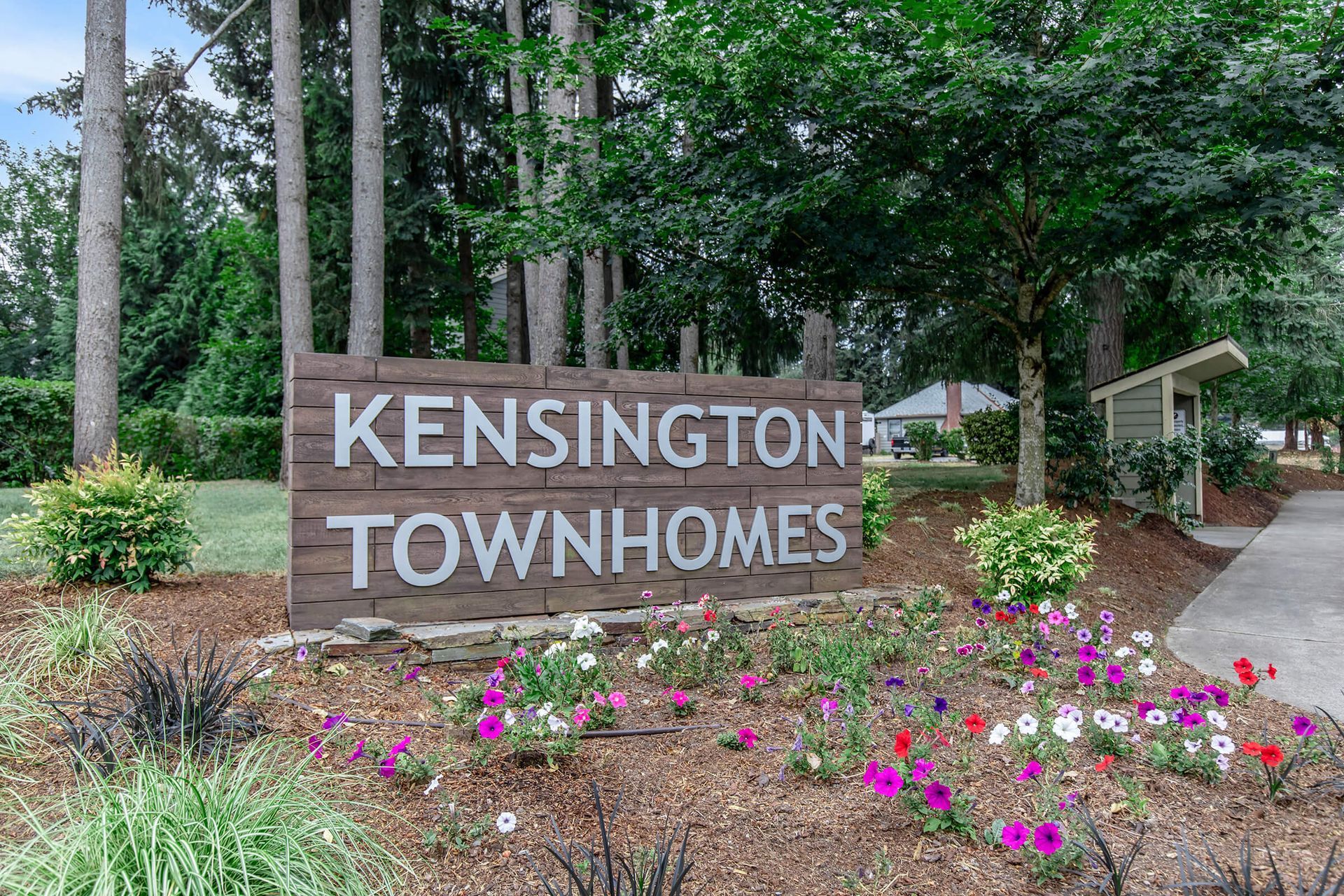 A sign for kensington townhomes is surrounded by flowers and trees.