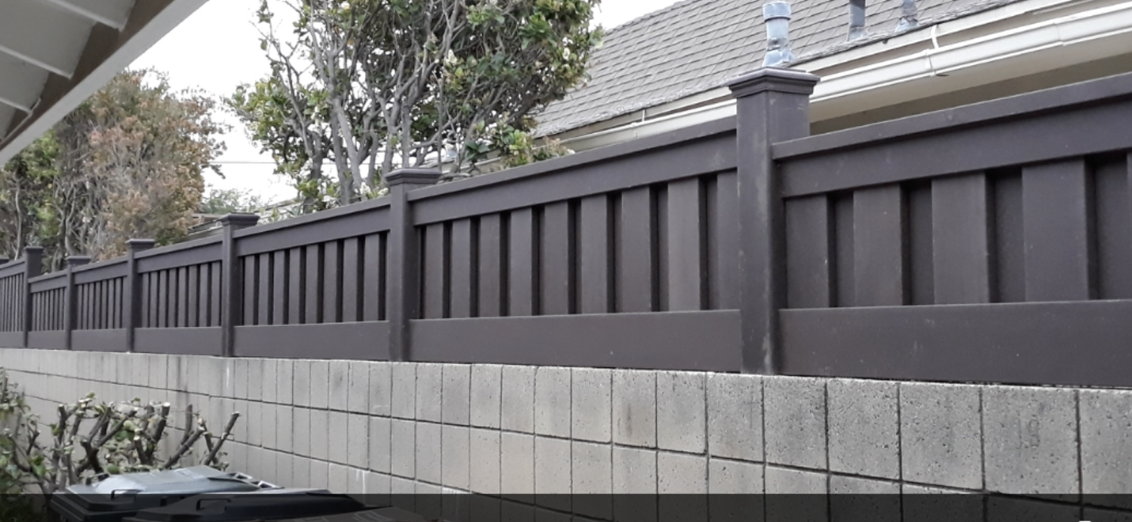 A dark gray fence with vertical slats sits on a concrete block wall, trees in the background.