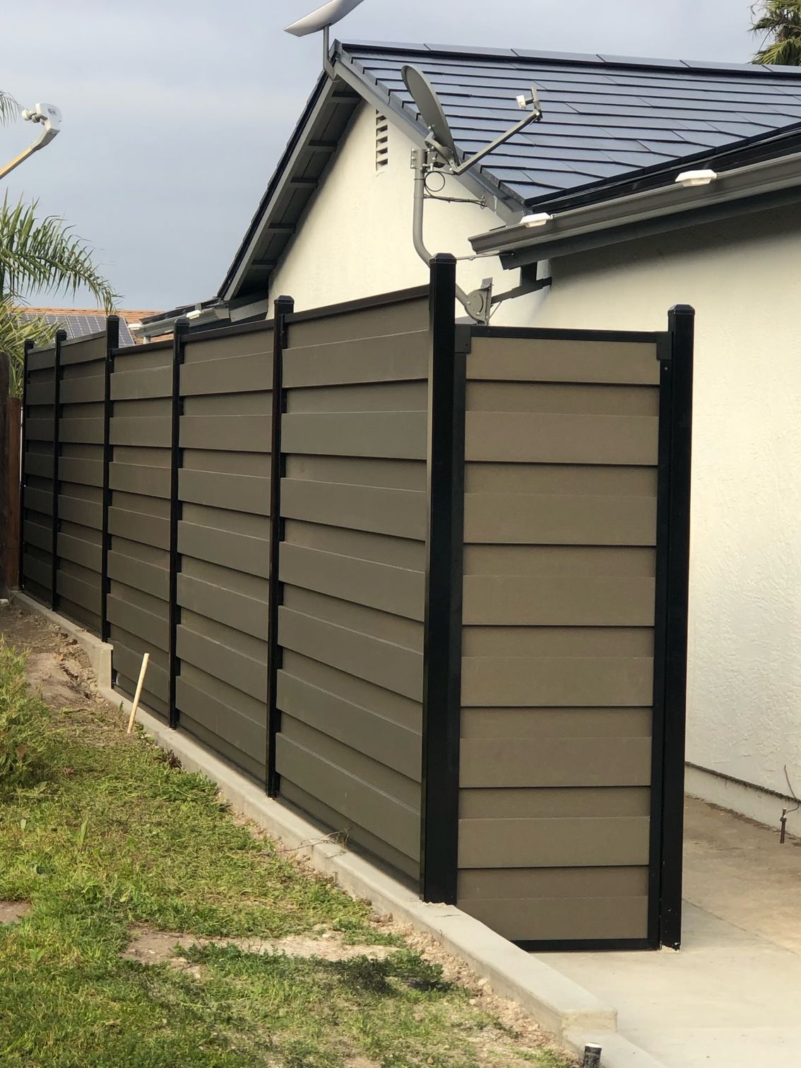 A brown and black horizontal slat fence runs along a grassy area next to a white house with a dark roof.