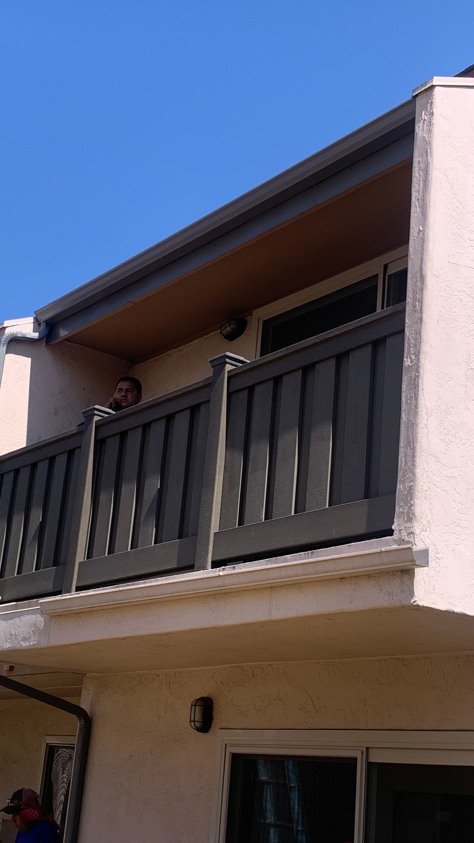 Man peeking over a gray balcony railing, beige building, blue sky.