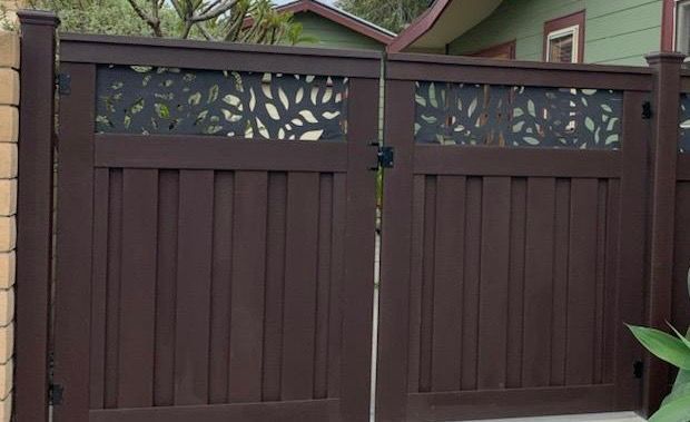 Brown wooden gate with leaf design, in front of a green house.