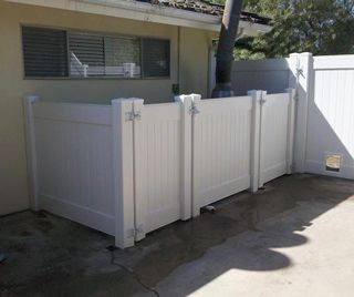 White vinyl privacy fence in front of green trees and dark soil.