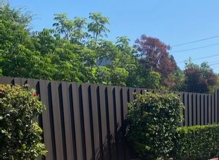 Brown vertical slat fence with green and reddish trees against a blue sky.