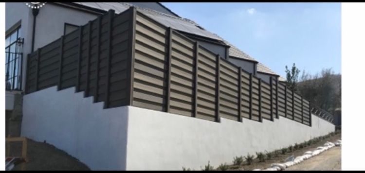 A long, dark fence atop a white wall, running along a sloping landscape beside a house.