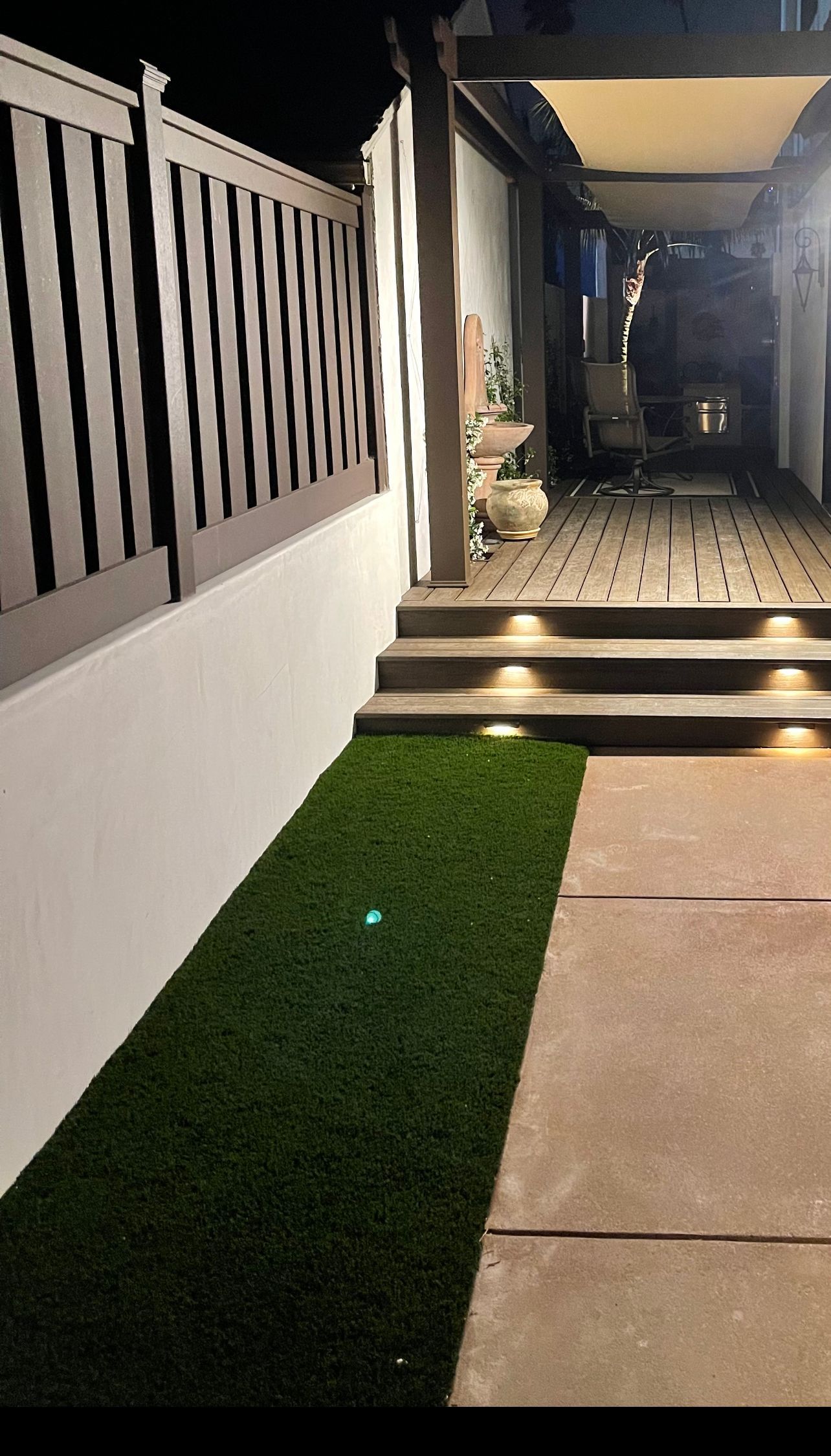 Narrow pathway with artificial turf, leading to a patio with steps, lit by recessed lights.