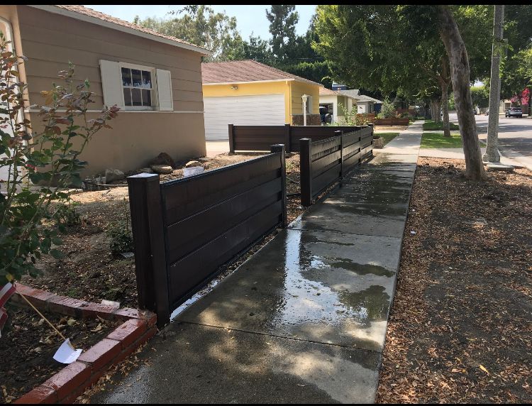Dark brown wooden fence along a sidewalk in front of a house, sunny day.
