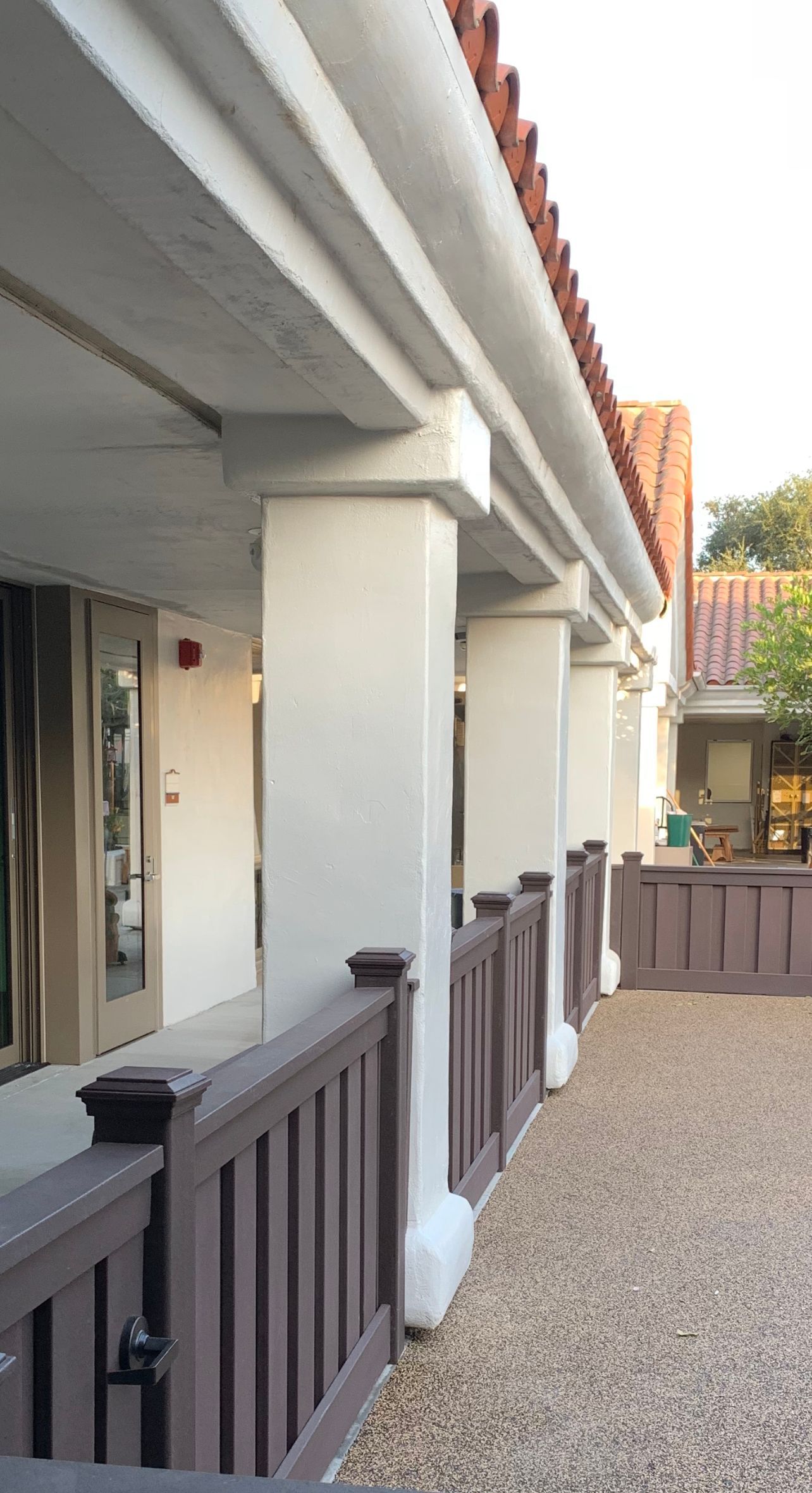 Covered walkway with white columns and brown railing; gravel ground.
