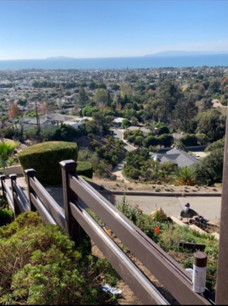 Brown fence overlooks a residential area, trees, and the ocean under a clear blue sky.