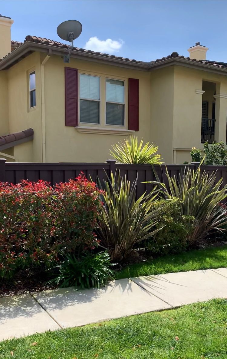 Two-story beige house with red shutters, brown fence, and various bushes along a sidewalk on a sunny day.