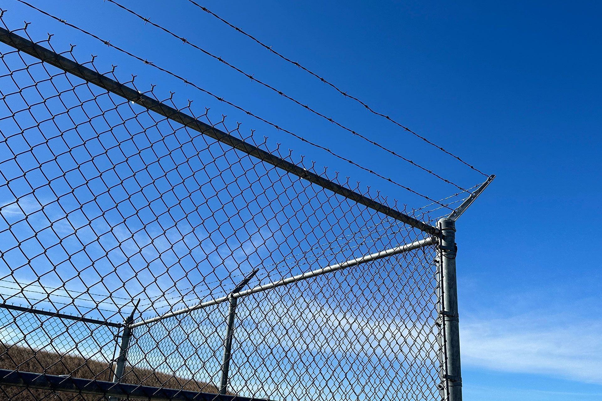 Chain-link fence with barbed wire against a bright blue sky.