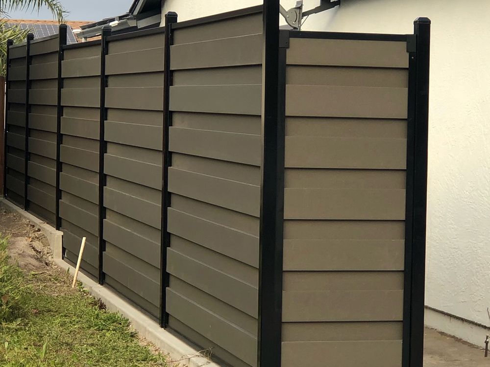 Dark-framed privacy fence with horizontal brown slats against a white building; a grassy area is on the left.