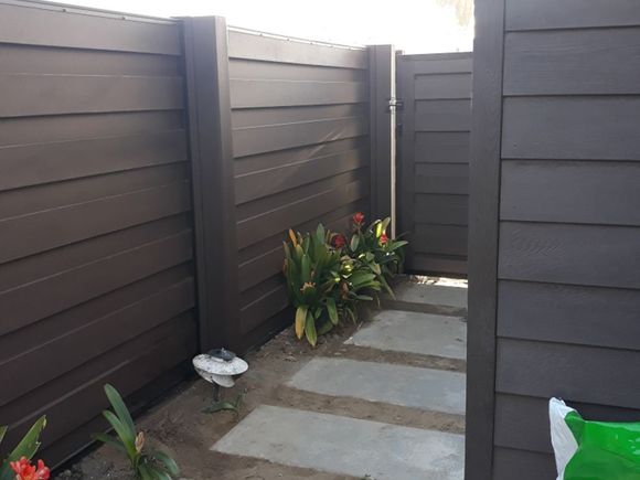 A dark brown privacy fence surrounds a stone pathway leading to a gate, with plants in between.