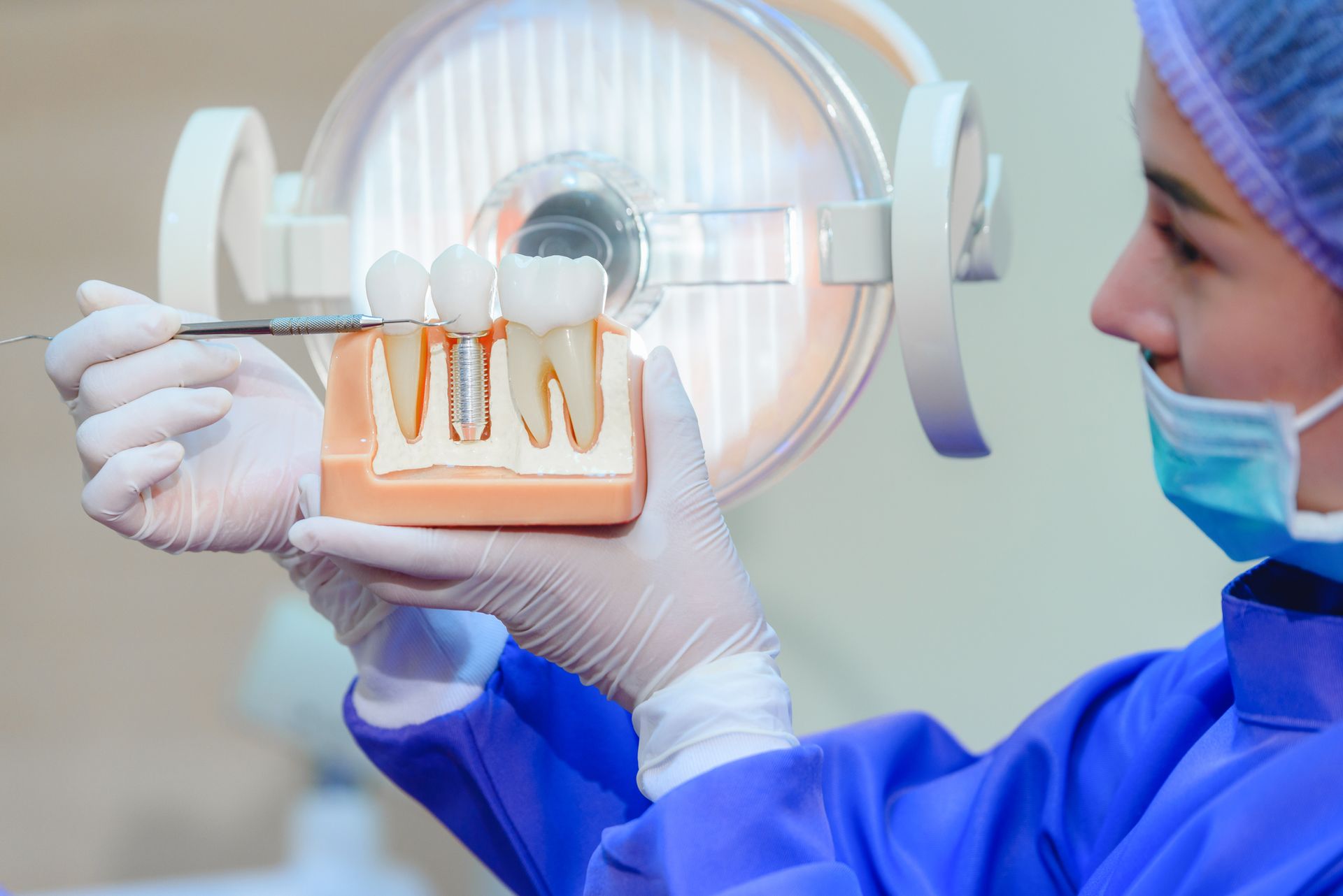 A dentist in a blue uniform, showing a tooth model and pointing to the crown of a dental implant.