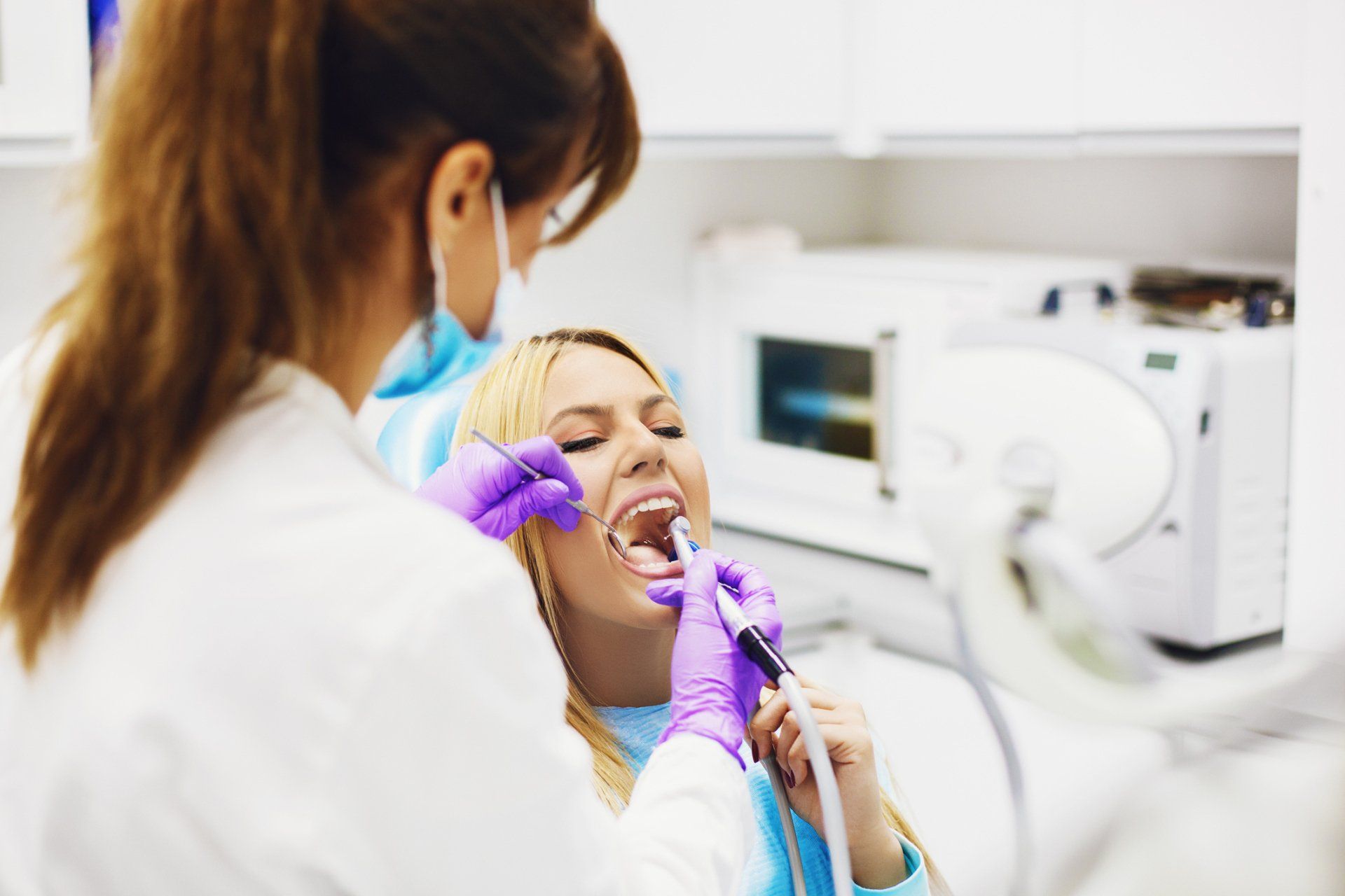 Dentist examining patient's teeth in a dental office. The patient has open mouth, while dentist holds tools and wears gloves.