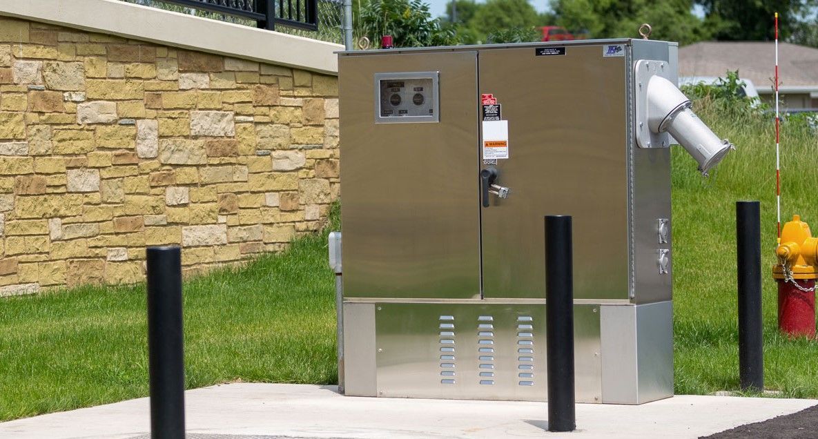 Metal utility box on concrete pad, black posts, fire hydrant, brick wall, grassy area.